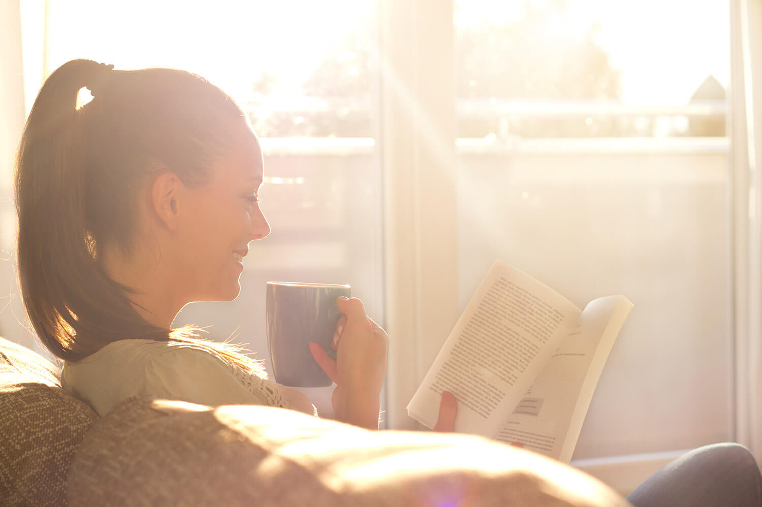 Frau genießt auf der Couch die Sonnenstrahlen, trinkt dabei Tee und ließt ein Buch.